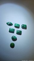 Seven green malachite cabochons arranged on a surface with light shadow, showing varied shapes including oval and rectangular with distinct banding patterns.