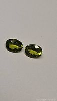 Pair of natural peridot gemstones with oval cut, bright apple green color, on a neutral background.