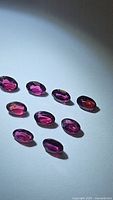 Ten oval cut purple-pink Rhodolite garnet gemstones laid flat on white background under bright light, showing color and clarity.
