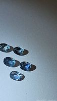 Five oval cut blue sapphire gemstones arranged on a flat neutral surface, showing overall color and clarity from top-down angle.
