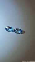 Pair of natural Swiss blue topaz oval cut gemstones placed side by side on a flat surface showing strong clarity and blue color.