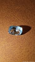 Close-up photo of a natural cushion cut aquamarine gemstone, blue color, laying on a textured brown surface showing its clarity and shape.