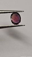 Natural oval cushion step cut sapphire held by gemstone tweezers showing pink orange color under light.