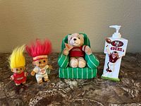 Four San Francisco 49ers themed decorative items arranged on a surface: two troll dolls, one teddy bear in a chair, and one lotion dispenser