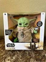 Front view of Star Wars The Mandalorian Baby Yoda toy inside original box, shows figure in beige robe holding silver ball accessory with included green frog accessory visible.
