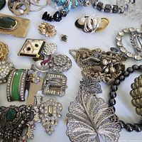 Close-up image of various vintage rhinestone and metal jewelry items including brooches, bracelets, and earrings in silver, gold, and green tones.