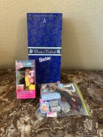 Photo showing one boxed Kelly Club doll named 'Amusement Park Nia', one Avon exclusive Barbie doll in closed blue Winter Velvet box, and miscellaneous small toy furniture items in a plastic bag on a surface.