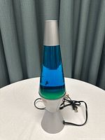 Blue lava lamp on white table with a dark curtain background and visible power cord.