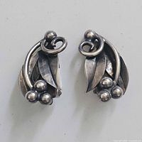Close-up photo of pair of sterling silver clip on earrings with leaf motif, swirl stems, and berry cluster details on white background.