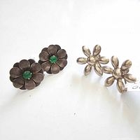 Two pairs of vintage silver flower-shaped screwback earrings on a white background: one with green glass center stones and another plain silver with rounded petals, showing tarnish and age wear.