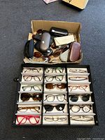 Overview of assorted eyeglasses, sunglasses, and cases in a cardboard box and a display tray