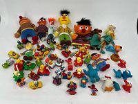 Full group of Sesame Street and Muppets themed toys and stuffed animals neatly arranged on white background with plush dolls in the back and smaller figures and vehicles in front.