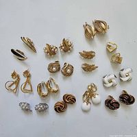 Overview of the 20 pair vintage Trifari goldtone clip-on earrings arranged on white background, showing various styles including faux pearl clusters, leaf motifs, enamel accents, and geometric shapes.