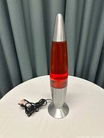 Red lava lamp on white table with green curtain background and power cord visible
