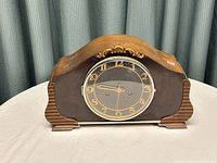 Front view of vintage wooden mantel clock showing curved wooden body, decorative floral motif, round gold-tone dial with Arabic numerals and clock hands, glass cover closed.