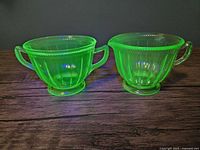 Pair of green uranium glass creamer and open sugar bowl in colonial fluted pattern illuminated under black light showing glowing effect and overall shape.