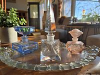 Front angled photo of Fostoria vanity tray displaying three vintage perfume bottles in blue, clear, and pink glass under natural light.