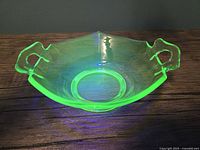 Hexagonal uranium glass dish glowing bright green under UV light, showing two handles and smooth finishing on wooden surface.