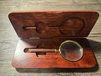 Magnifying glass inside an open cherry wood box showing the lens and handle with brass accents fitted into carved inset.