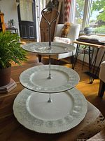 Three-tiered fine bone china dessert stand with floral decoration and metal handle, displayed in a living room setting on a wooden table.