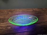 Oval uranium glass dish glowing bright green under UV light on wooden surface.