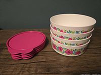 Stack of five white Tupperware bowls with colorful floral designs on the sides, arranged next to a stack of five bright pink lids.