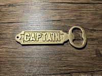 Top view of the brass bottle opener with 'CAPTAIN' embossed on it against wooden background.