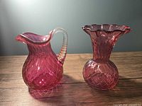 Photo of vintage cranberry glass pitcher and vase on wood surface against plain wall, showing their shape and color