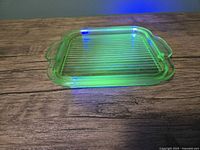 Top and side view of the square uranium glass tray glowing under UV light, showing ribbed pattern and integrated handles.