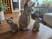 Three porcelain cats (one adult and two kittens) on a wooden surface with indoor background