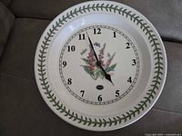 Full view of the round ceramic clock face showing the foxglove flower illustration, green leaf border, black hour and minute hands, and Arabic numerals.