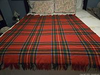 Full view of the vintage Scottish tartan throw laid on a bed showing the red dominant tartan pattern with green, yellow, white and blue stripes and fringed edges.
