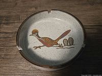 Top view of speckled ceramic ashtray with roadrunner and cactus design in center, showing cigarette rests on rim.