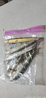 Five large diving fishing lures of various metallic colors inside a clear plastic zip bag.