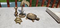 Photo of five brass decorative items on an outdoor table including a bird sculpture, two turtles, a candlestick holder, and a turtle-shaped dish