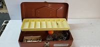 Open brown metal tackle box showing cream-colored compartment tray and assorted fishing tackle including line and bobber.