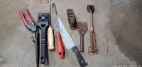 Photo showcasing six hand tools including pliers, paint scrapers, knife, and rusted scrapers arranged horizontally on a concrete floor.