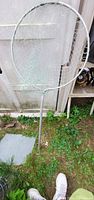 Large fishing net propped against an outdoor wall in grass, showing circular metal frame painted white and attached mesh netting. Metal handle extends downward into grass.