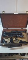 Open brown lined case containing a large black tactical-style hunting scope with dials and multiple small accessories including springs, lens covers, and adjustment rings.