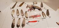 Photo showing pile of 19 assorted metal fishing spoon lures on white background, some tarnished, some painted, some in plastic packaging.