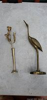 Photo of tall brass bird figurine and brass flower vase sculpture on a light grey surface