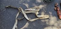 Two beige tie down straps with metal ratchet buckles lay unfolded on asphalt next to a rusty metal chain load binder with hooks on both ends.