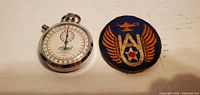 Metal mechanical stopwatch and a circular embroidered arm patch side by side on a white surface.