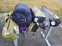 Overview of five bagged camping items on stand: one olive tent bag, four assorted sleeping bag sacks