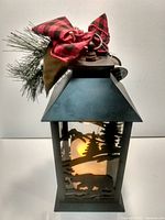 Front view of lantern showing cut-out scene, LED candle glow, greenery and bow