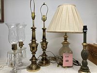 Six assorted table lamps on a wooden surface showing detailed view of brass trophy urn lamps, ceramic lamp with pleated shade, green pineapple motif lamp, and crystal hurricane lamps with glass shades.
