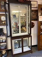 Full front view of the antique display cabinet showing glass doors on upper and lower sections with mahogany frame and side panels. Doors are open to reveal interior glass shelves.