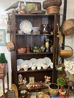 Front view of tall antique wooden shelf filled with various decorative items including plates, pitchers, vases, baskets, and figurines, showing shelf design and wood finish.