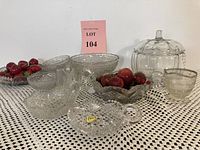 Full lot of clear decorative glass items, including lidded cookie jar, pitcher, bowls, and candle holder displayed on crocheted cloth with decorative fruit inside some bowls.