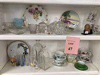 Full view of shelf showing assortment of porcelain plates with floral designs, teacups with saucers, clear glass serving pieces, decorative bottles, and a commemorative medal.